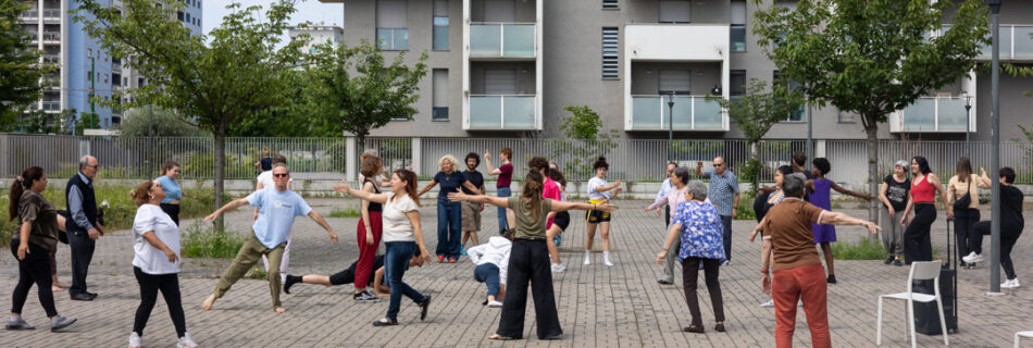 Un nutrito gruppo intergenerazionale si muove nello spazio pubblico all’aperto durante il laboratorio “Dance Well”.
