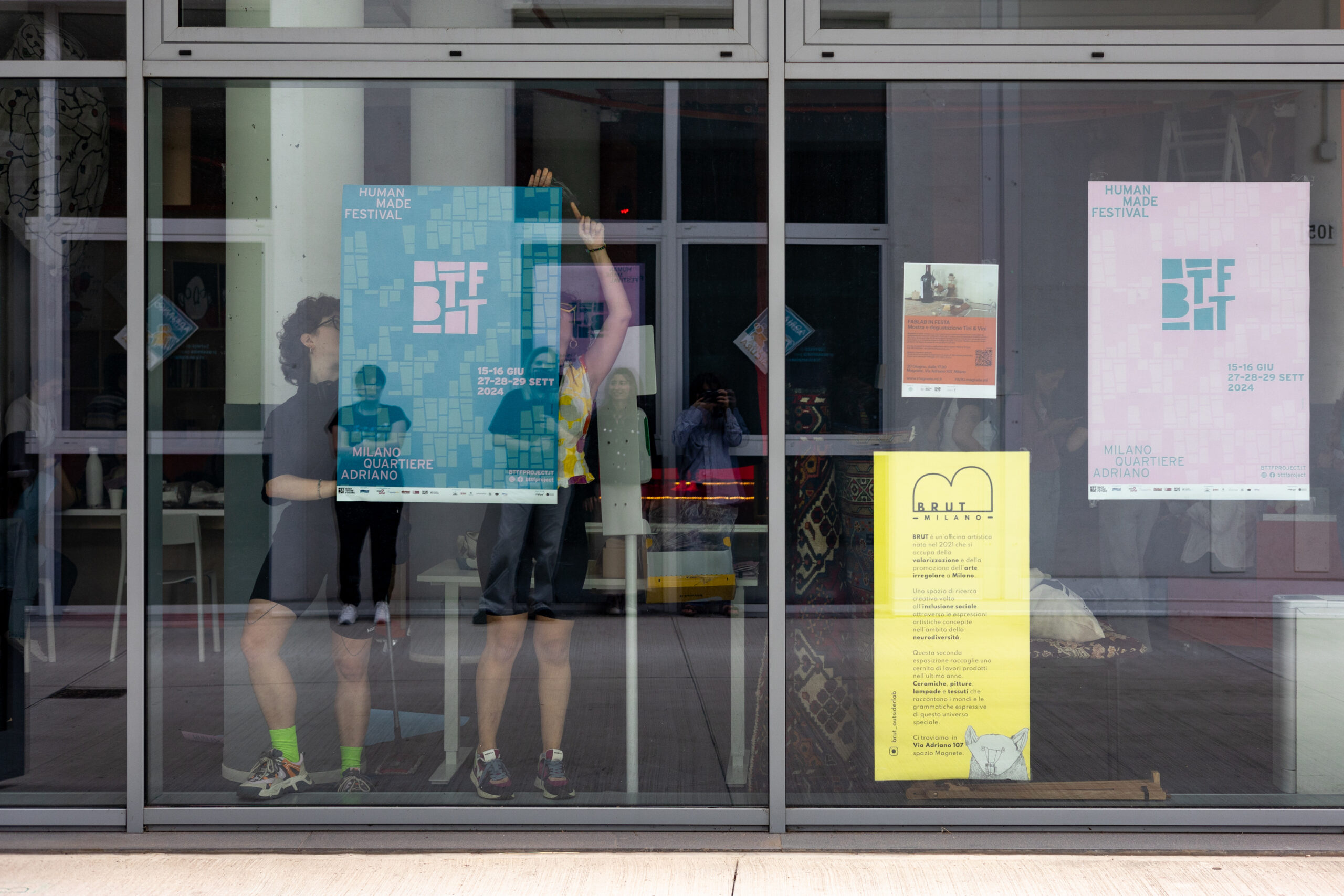 Due ragazze della Direzione Artistica Partecipata Under 30 attaccano un poster dietro una vetrata.