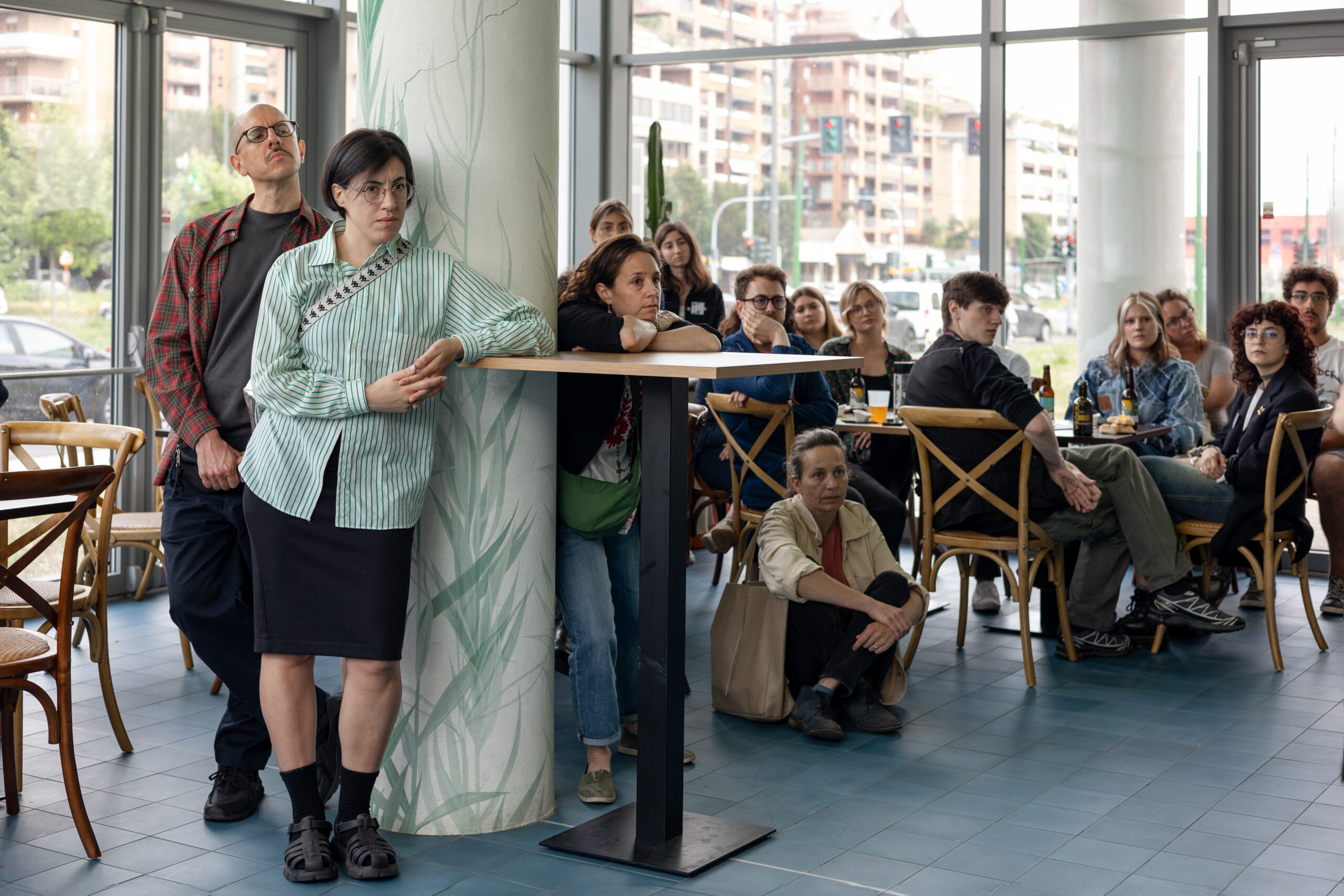 Alcune persone in piedi e sedute in un bistrot ascoltano qualcuno parlare.