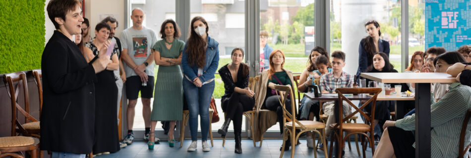 Alcune persone sedute e in piedi ascoltano una donna dello staff con i capelli corti mentre inaugura il festival con un discorso di apertura.