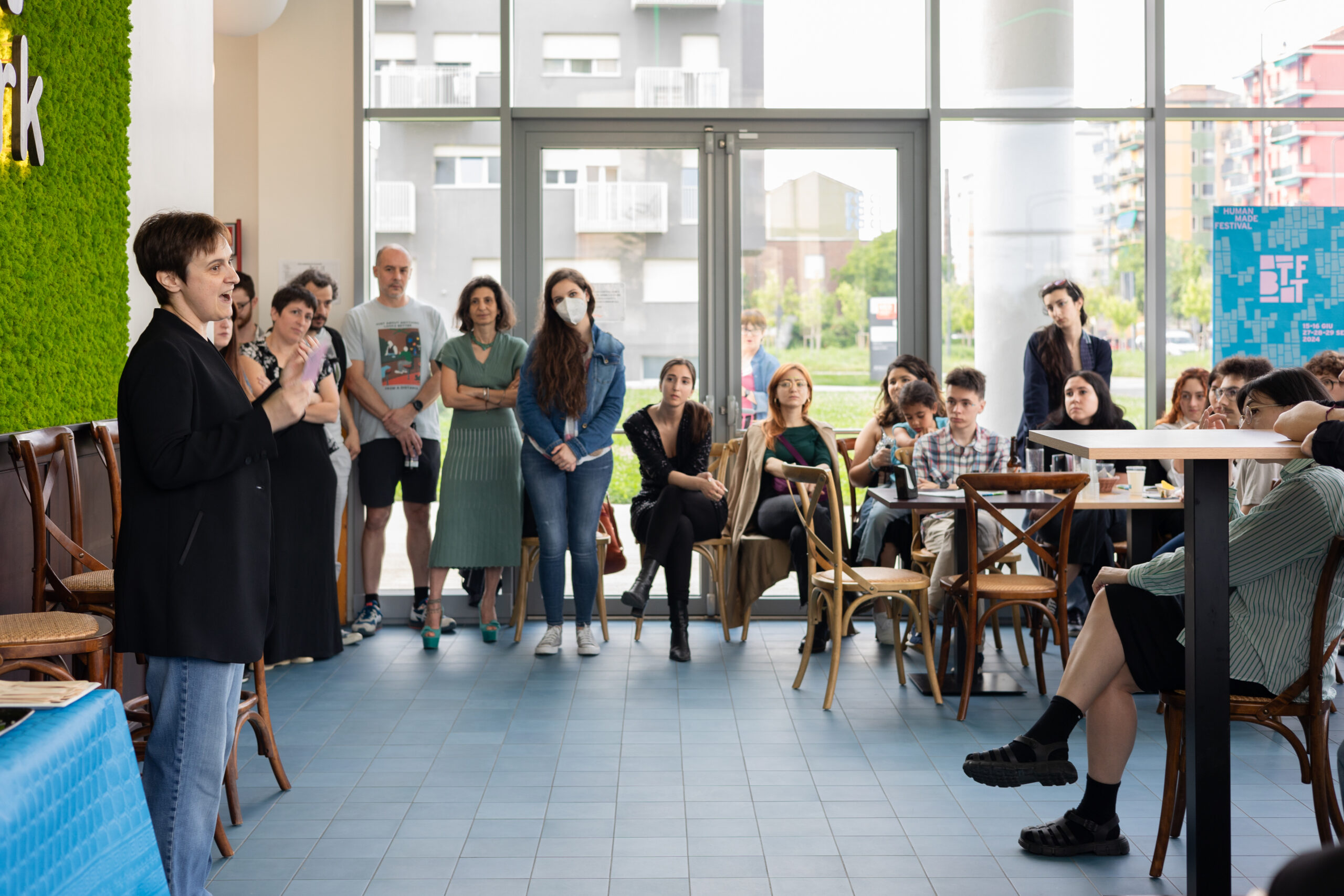 Alcune persone sedute e in piedi ascoltano una donna dello staff con i capelli corti mentre inaugura il festival con un discorso di apertura.