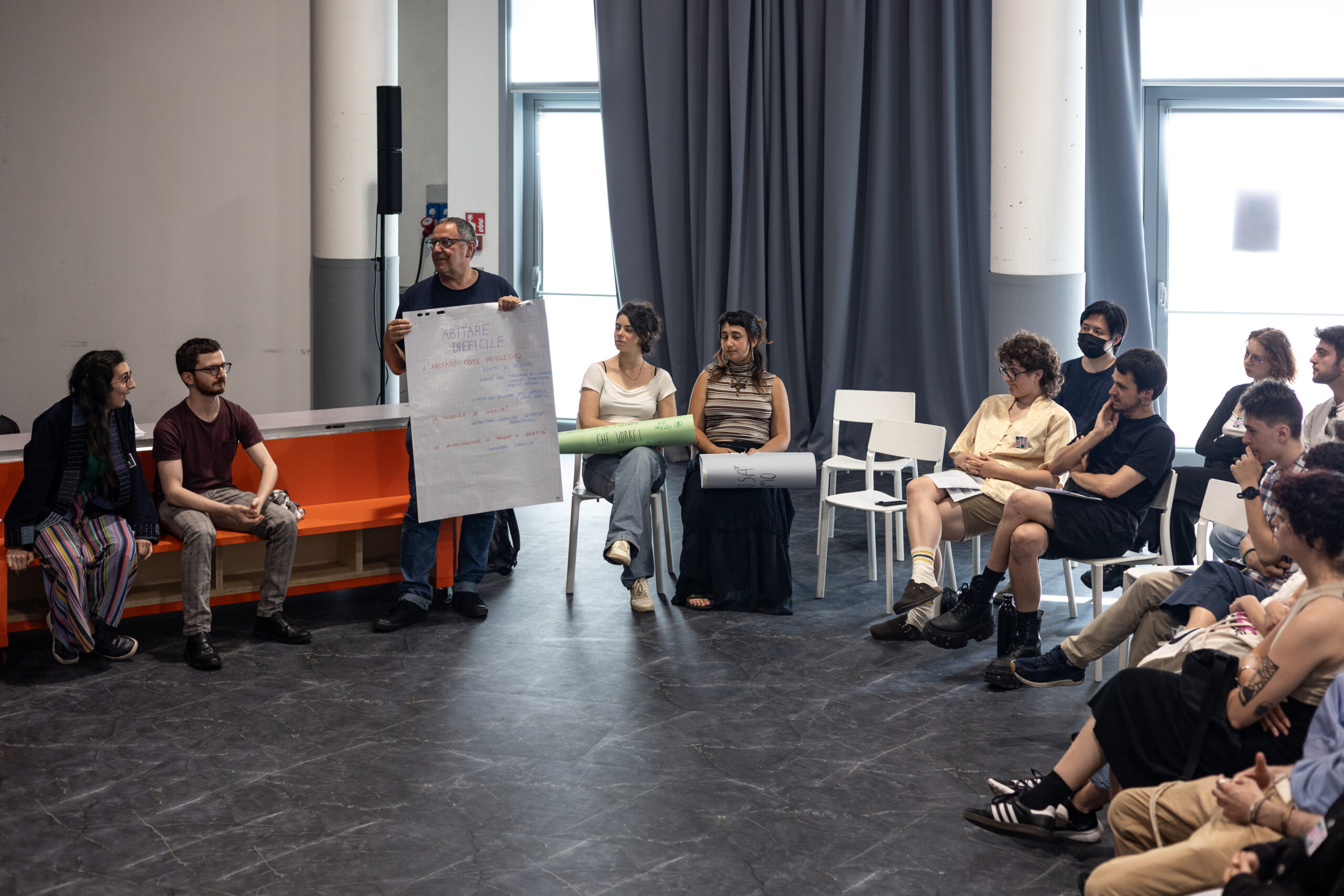 Un relatore del laboratorio “Esercizi di immaginazione collettiva”, con un cartellone in mano, spiega mentre delle persone in cerchio ascoltano.