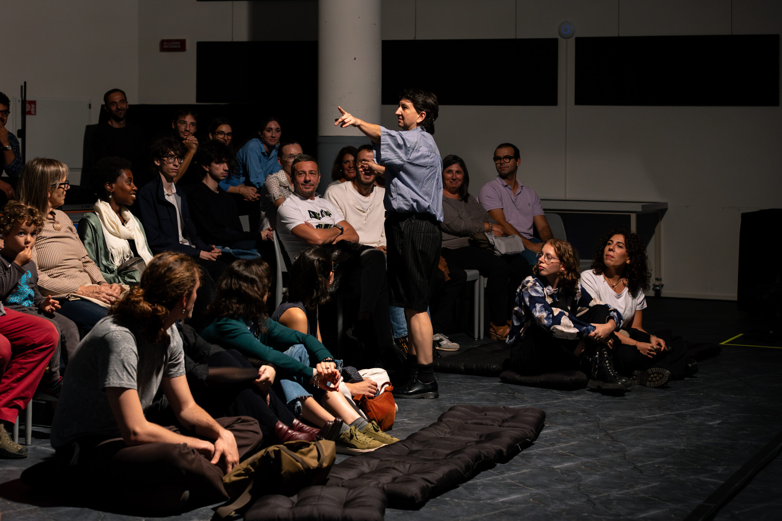 Il pubblico seduto osserva la performer di “Insectum” in Quartiere Adriano indicare qualcosa.