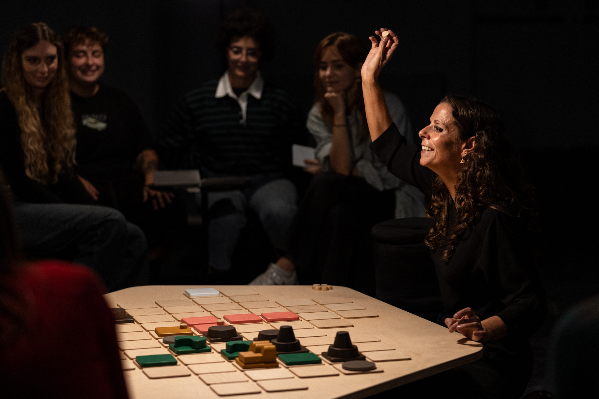 La performer di “Eutopia” tiene il braccio alzato con una pedina del gioco che ha sul tavolo di fronte, sullo sfondo alcune persone del pubblico sorridono.