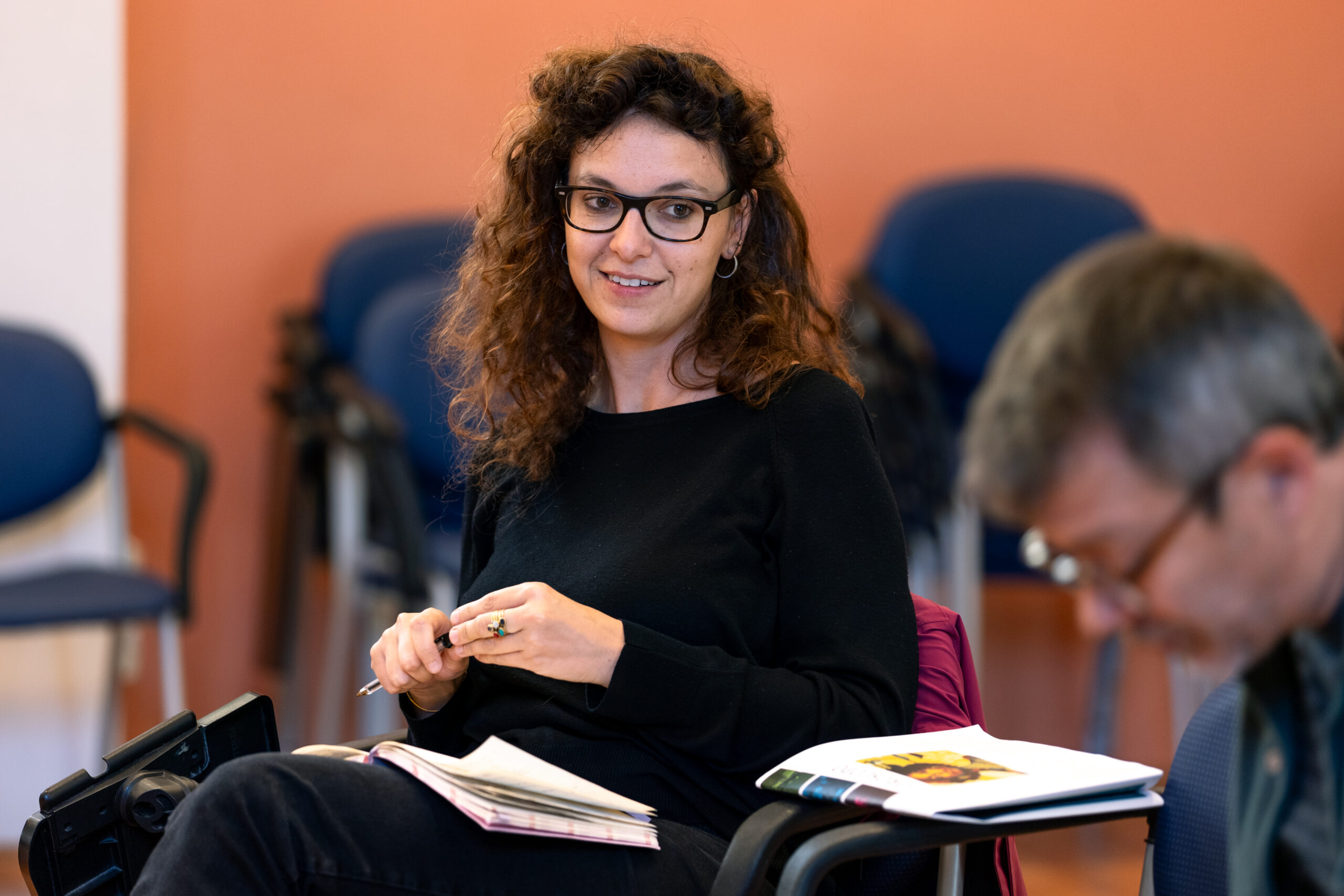 La moderatrice del convegno “InVisible”, una giovane donna con gli occhiali scuri e i capelli ricci castani, sorride e prende appunti su un taccuino