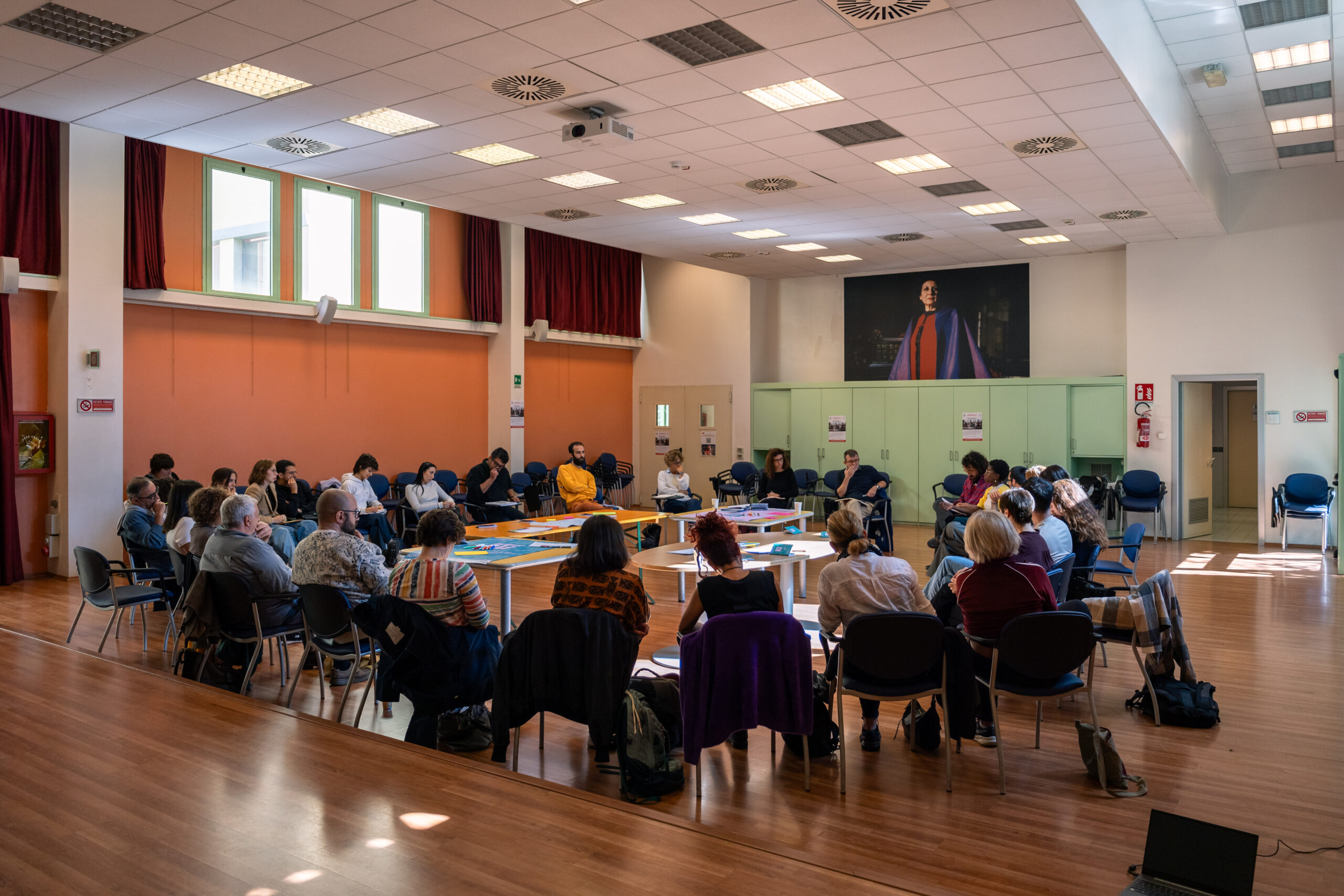 Trenta partecipanti al convegno “InVisible” sono seduti circolarmente per iniziare il dibattito sulla sostenibilità nell’auditorium di Casa della Carità.